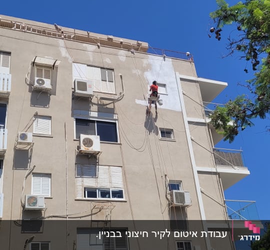 אדם צובע קיר חיצוני של בניין גבוה בעזרת חבלים.